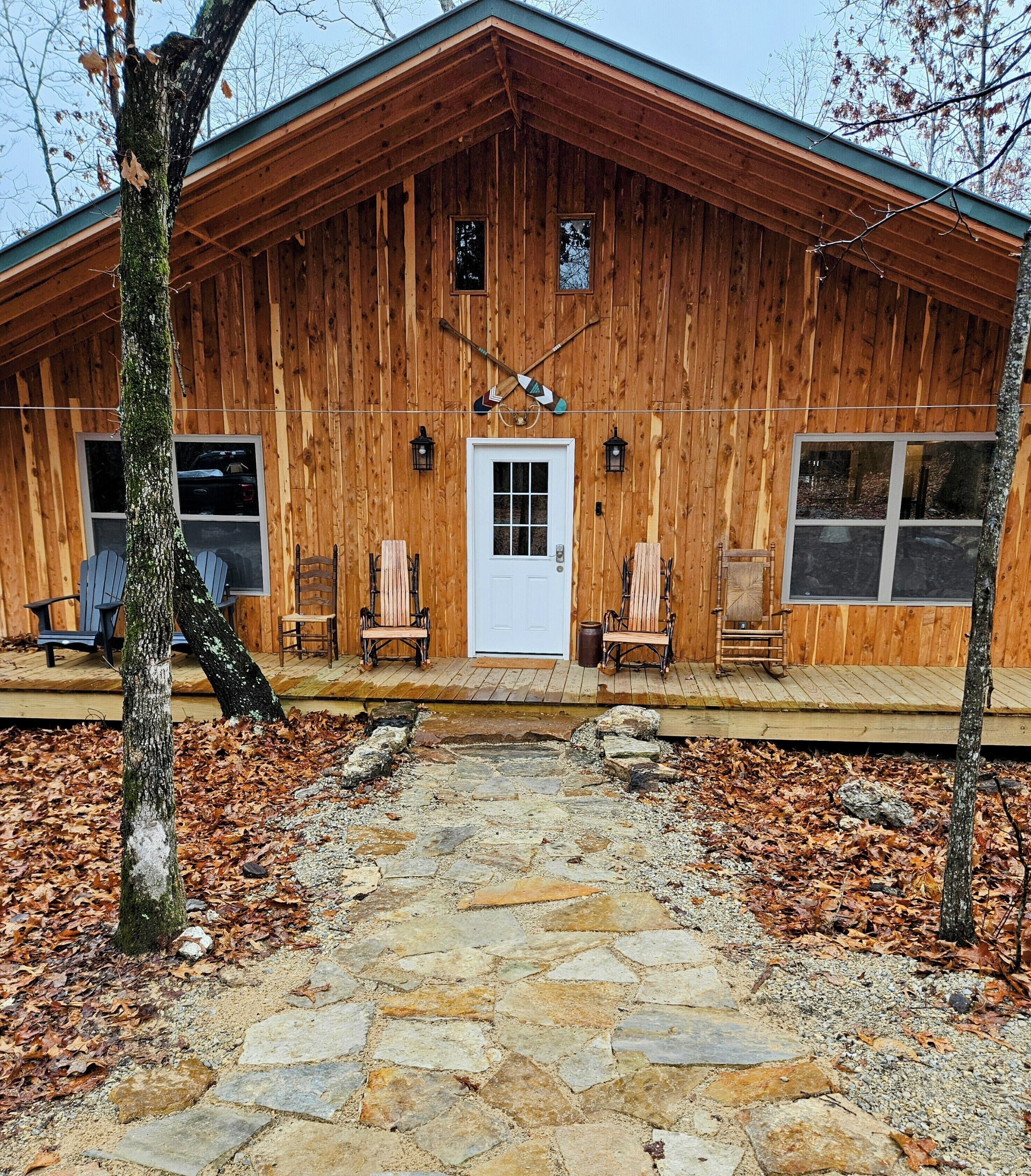 A wooden cabin with a stone pathway leading to the entrance, surrounded by trees and autumn leaves.