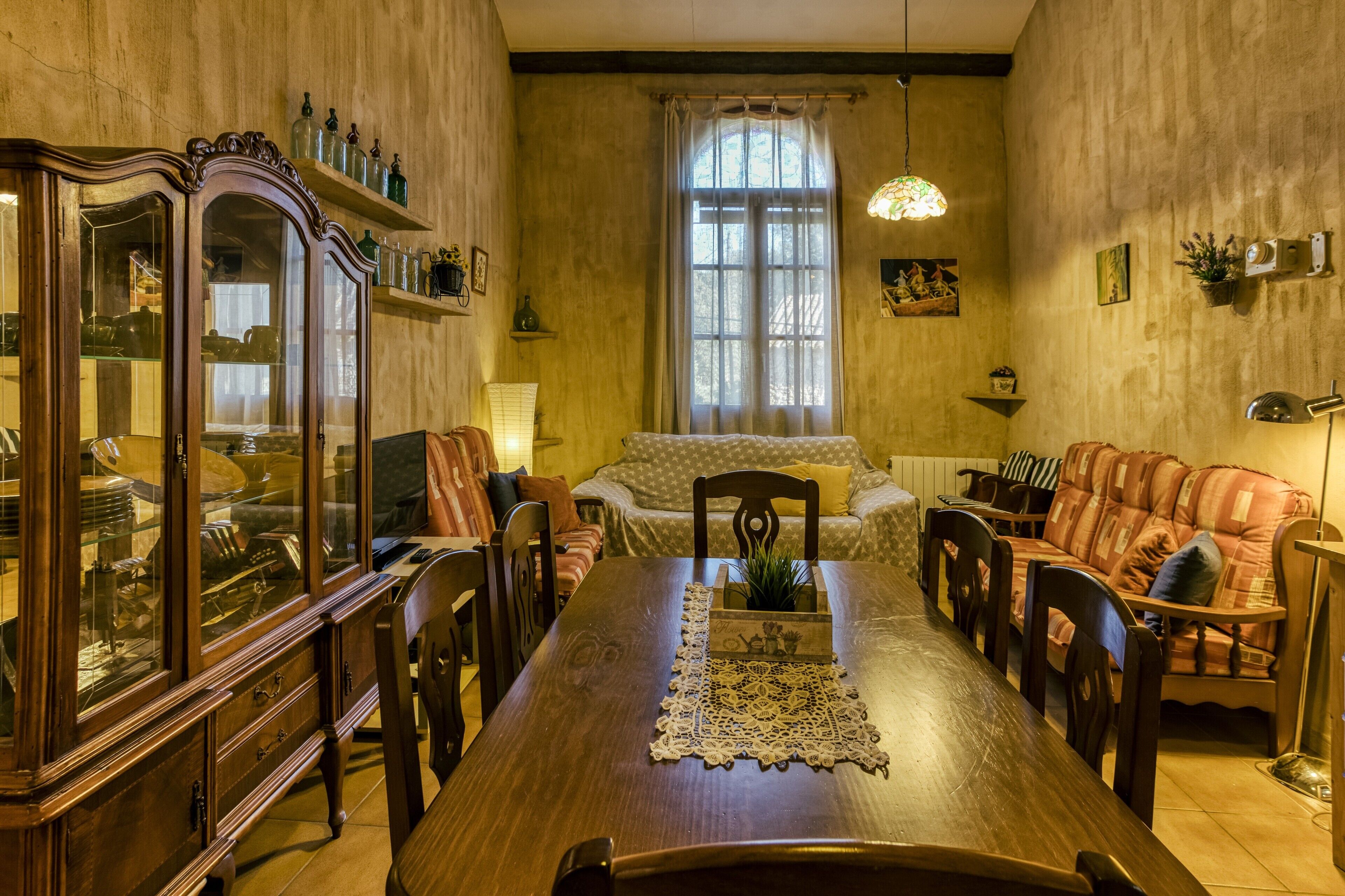 Un comedor con una mesa de madera, sillas, un aparador con puertas de cristal, un sofá y una ventana con cortinas.