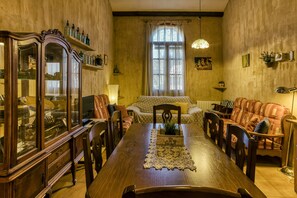 A dining room with a wooden table, chairs, a cabinet with glass doors, a sofa, and a window with curtains.