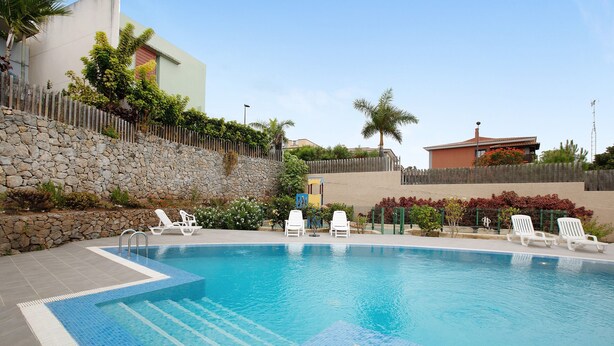 A swimming pool with lounge chairs, a stone wall, and a wooden fence. - Vacation Home 'La Quinta Adosado' with Sea View, Shared Pool & Wi-Fi (Santa Ursula)