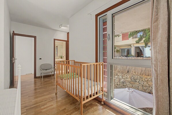 A room with a wooden crib, a chair, and a large window with a view of a building and stone wall.