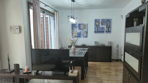 A modern living room with a flat-screen TV, dining table, and chairs. There's a kitchen area with cabinets and a refrigerator. - Vacation Home 'La Quinta Adosado' with Sea View, Shared Pool & Wi-Fi (Santa Ursula)