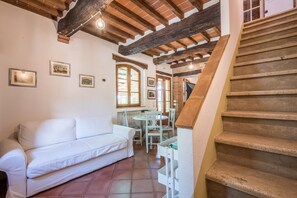 A room with a white sofa, a dining table with chairs, a staircase, and wooden beams on the ceiling.