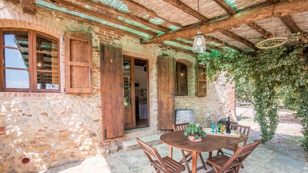 Un bâtiment en pierre avec des volets en bois, une table ronde en bois entourée de chaises et un luminaire suspendu.