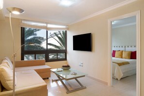 A modern hotel room with a sofa, a glass coffee table, a flat-screen TV, and a bed with red and gold bedding.