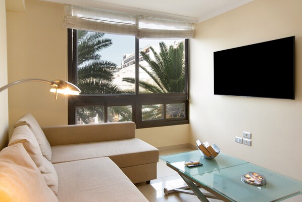 A modern living room with a sofa, a glass coffee table, a flat-screen TV, and a window with a view of palm trees.