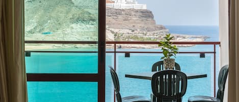 A balcony with a table and chairs, overlooking a coastal view with a cliff and a building.