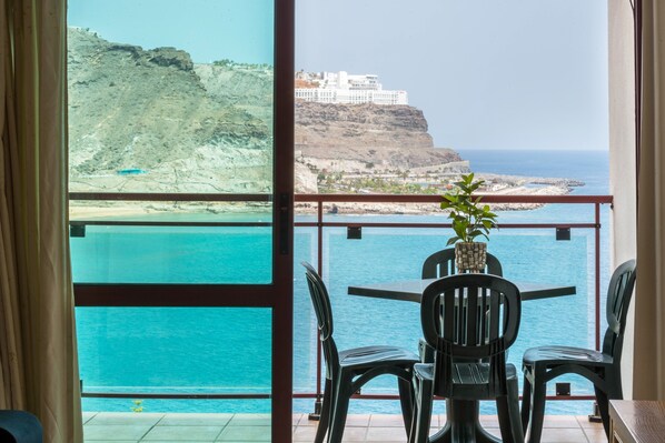 A balcony with a table and chairs, overlooking a coastal view with a cliff and a building.
