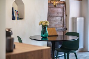 Une table ronde surmontée d’un vase turquoise contenant des fleurs blanches, une chaise verte et une bibliothèque remplie de livres, le tout dans une pièce dotée d’une porte en bois rustique et d’un radiateur.
