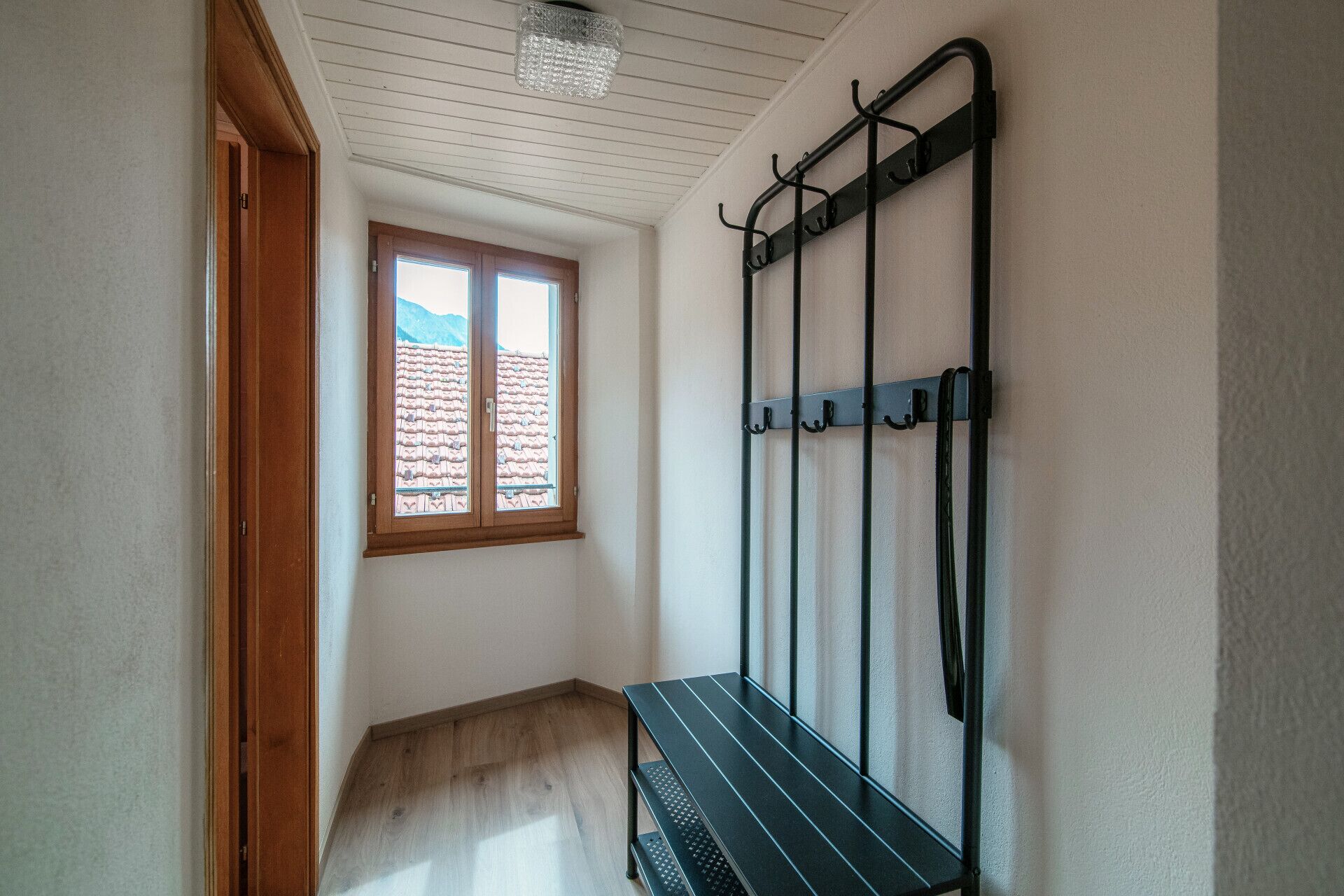 A room with a wooden bench, a metal rack with hooks, and a window with a view of a tiled roof.