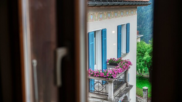 Une vue à travers une porte ouverte d’un bâtiment aux volets bleus, doté d’un toit en tuiles et d’un balcon orné de fleurs roses.