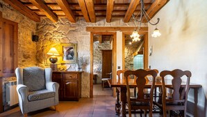 Un interior rústico con paredes de piedra vista, vigas de madera, una zona de comedor con sillas y una mesa de madera, un sillón estampado y una lámpara.
