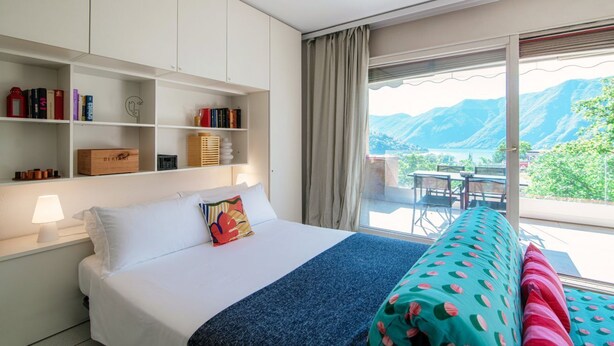 A hotel room with a bed, a bookshelf, a desk, and a view of mountains. - Open-space apartment with lake view (PANORAMIC STUDIO) (Lugano)