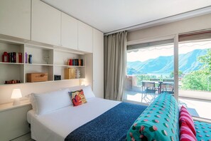 A hotel room with a bed, a bookshelf, a desk, and a view of mountains.