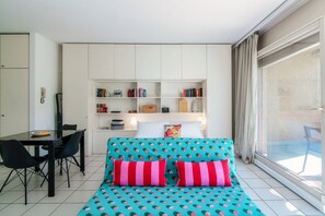 A modern room with a teal patterned bed, a white bookshelf, a dining area with a table and chairs, and a large window with curtains.