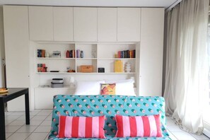 A room with a teal sofa with pink and white stripes, a white bookshelf filled with books and decorative items, and a window with curtains. - Open-space apartment with lake view (PANORAMIC STUDIO) (Lugano)
