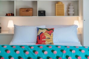 A neatly arranged hotel room with a bed, pillows, a nightstand, and a shelf with various items.