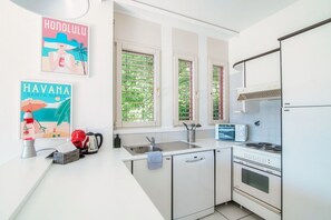 A modern kitchen with white cabinets, a sink, a stove, and a microwave. There are two windows with blinds, and a poster of Havana on the wall. - Open-space apartment with lake view (PANORAMIC STUDIO) (Lugano)