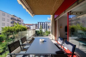 A balcony with a table and chairs, a glass railing, and a view of other buildings and greenery.