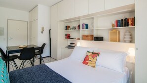A hotel room with a bed, a desk with chairs, and a bookshelf with books and decorative items. - Open-space apartment with lake view (PANORAMIC STUDIO) (Lugano)