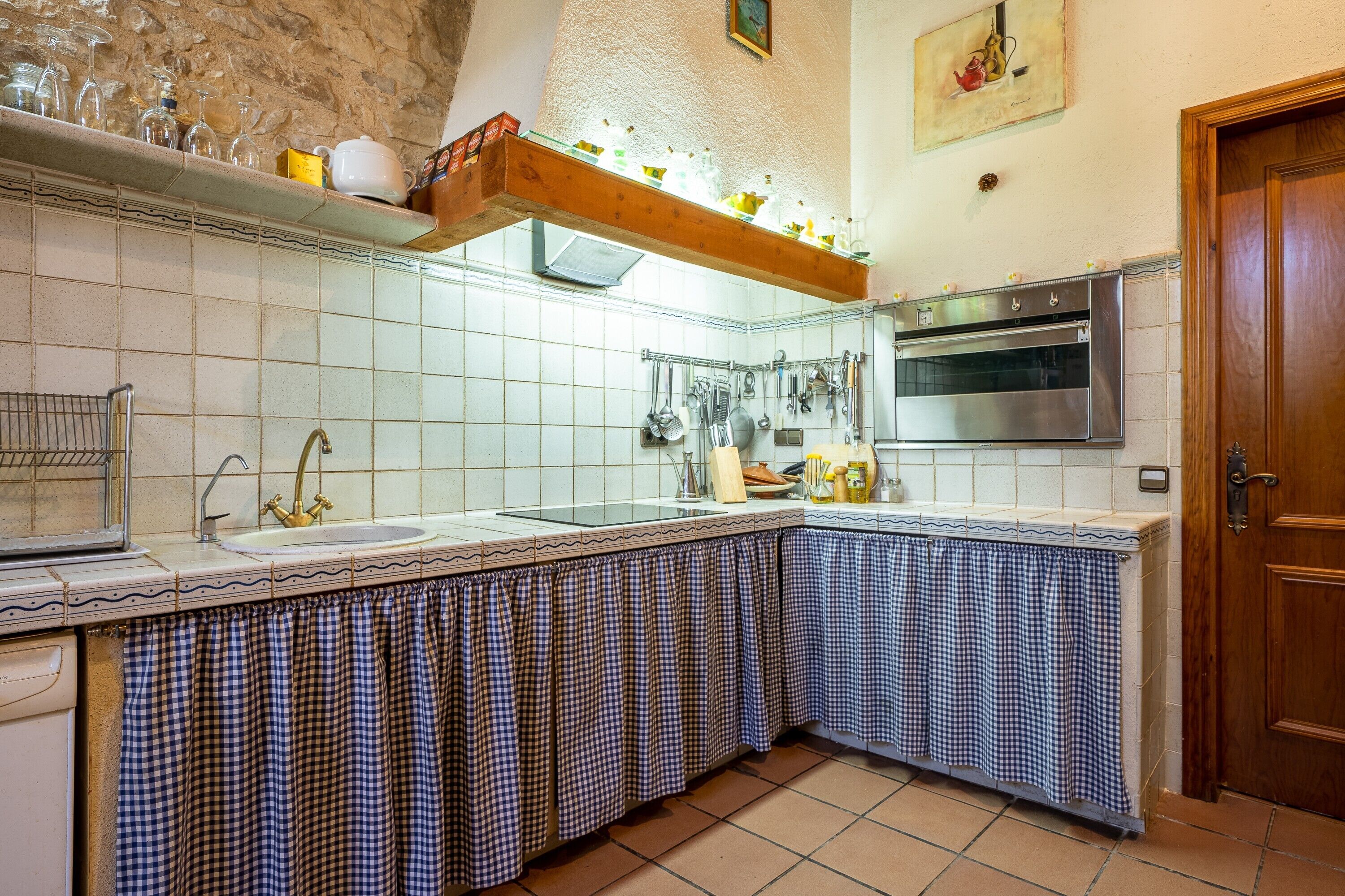 Una cocina con salpicadero de azulejos, una estantería de madera con cristalería, un horno de acero inoxidable y un mantel a cuadros cubriendo la encimera.