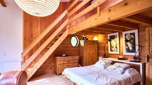 Une chambre dans une cabane en bois, avec un espace de couchage en mezzanine, une commode en bois, un bureau avec une lampe et une fenêtre ronde.