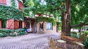 Une cour pavée de pierres, avec un bâtiment rustique, un arbre et une table en métal entourée de chaises.