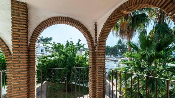 En balkon med murarkader, gelænder i metal og udsigt til grønne områder og bygninger.