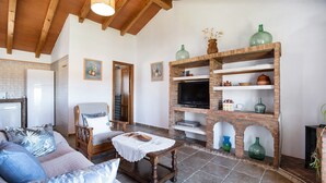 A living room with a sofa, armchair, wooden coffee table, TV, brick fireplace, and tiled floor. - Holiday Home 'Casa Andrea' with Mountain Views, Shared Pool & Wi-Fi (Barbate)