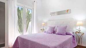 A bedroom with a large bed, two bedside tables, a window with a view of a cactus, and a wall-mounted light fixture.