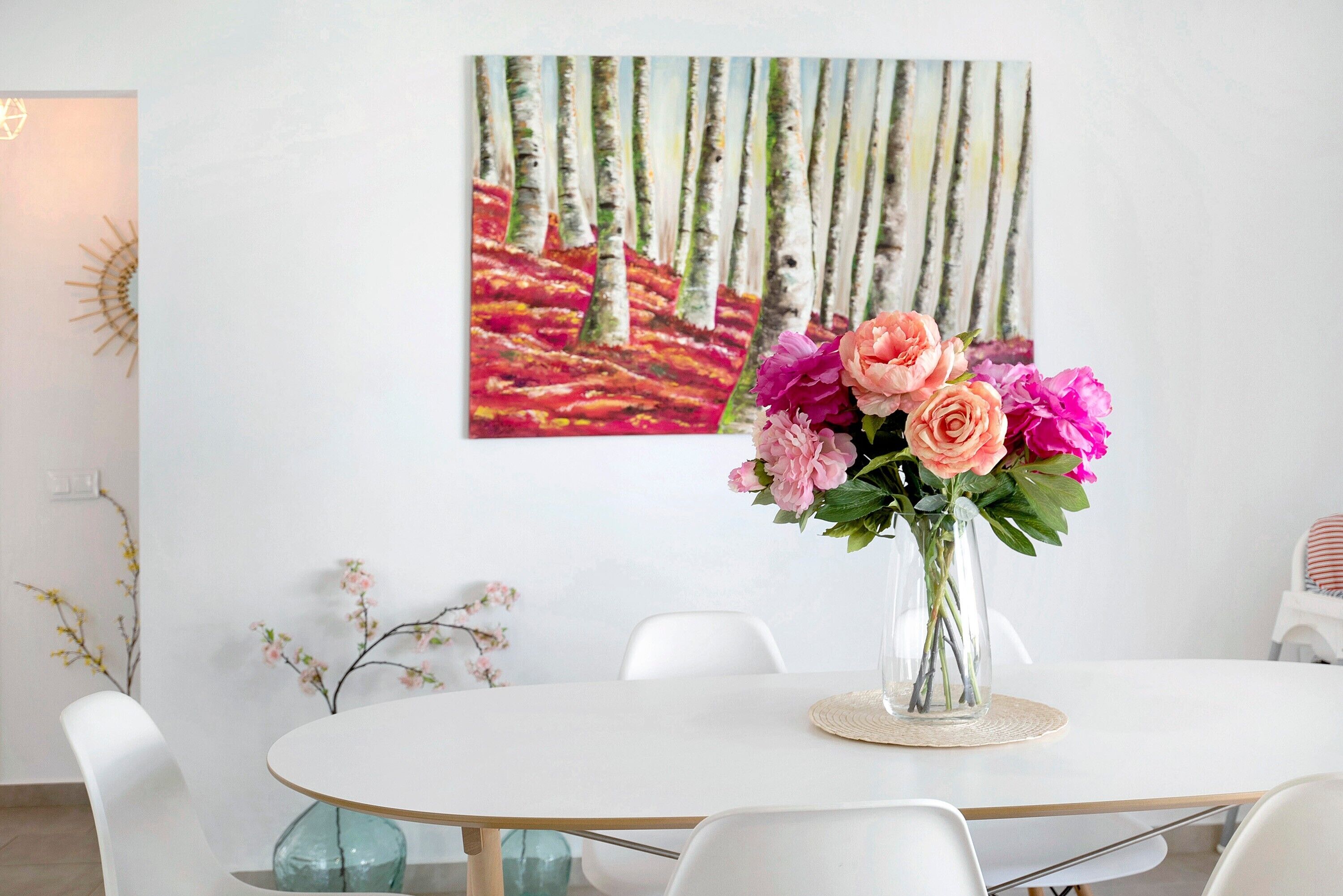 A dining area with a white table, clear glass vase with flowers, and a painting of birch trees on the wall.