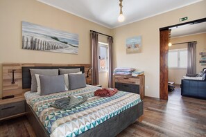 Un dormitorio con cama, mesita de noche, cabecera de madera, un cuadro, ventana con cortinas, suelo de madera y una puerta que da a otra habitación.