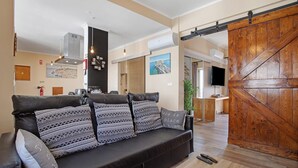 Een moderne woonkamer met een zwarte leren bank, gestreepte kussens, een schuifdeur in boerderstijl, en een keuken op de achtergrond.