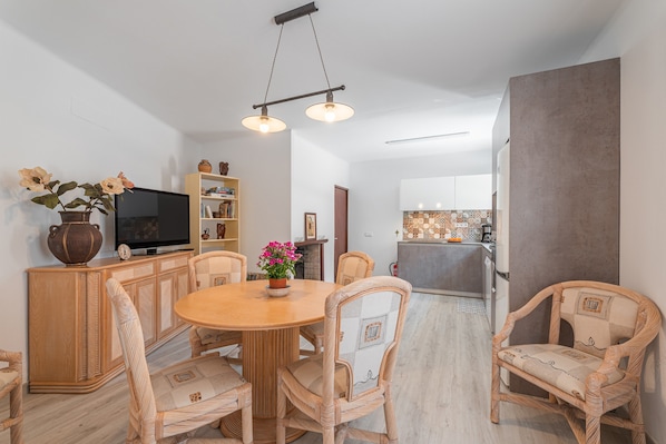 Un comedor con sillas de mimbre, una mesa redonda de madera, un televisor sobre un mueble y una cocina al fondo.