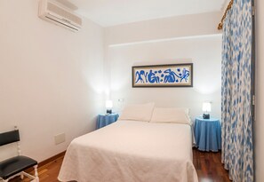 Une chambre à coucher comprenant un lit, deux tables de chevet avec des lampes, une chaise, un climatiseur et une œuvre d’art accrochée au mur.