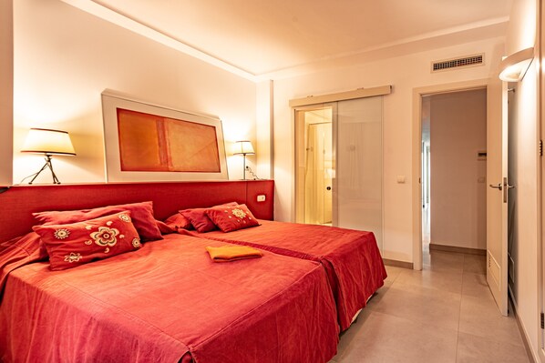 A hotel room with a large bed, two bedside tables, a wall-mounted artwork, a door leading to another room, and a light fixture on the ceiling.