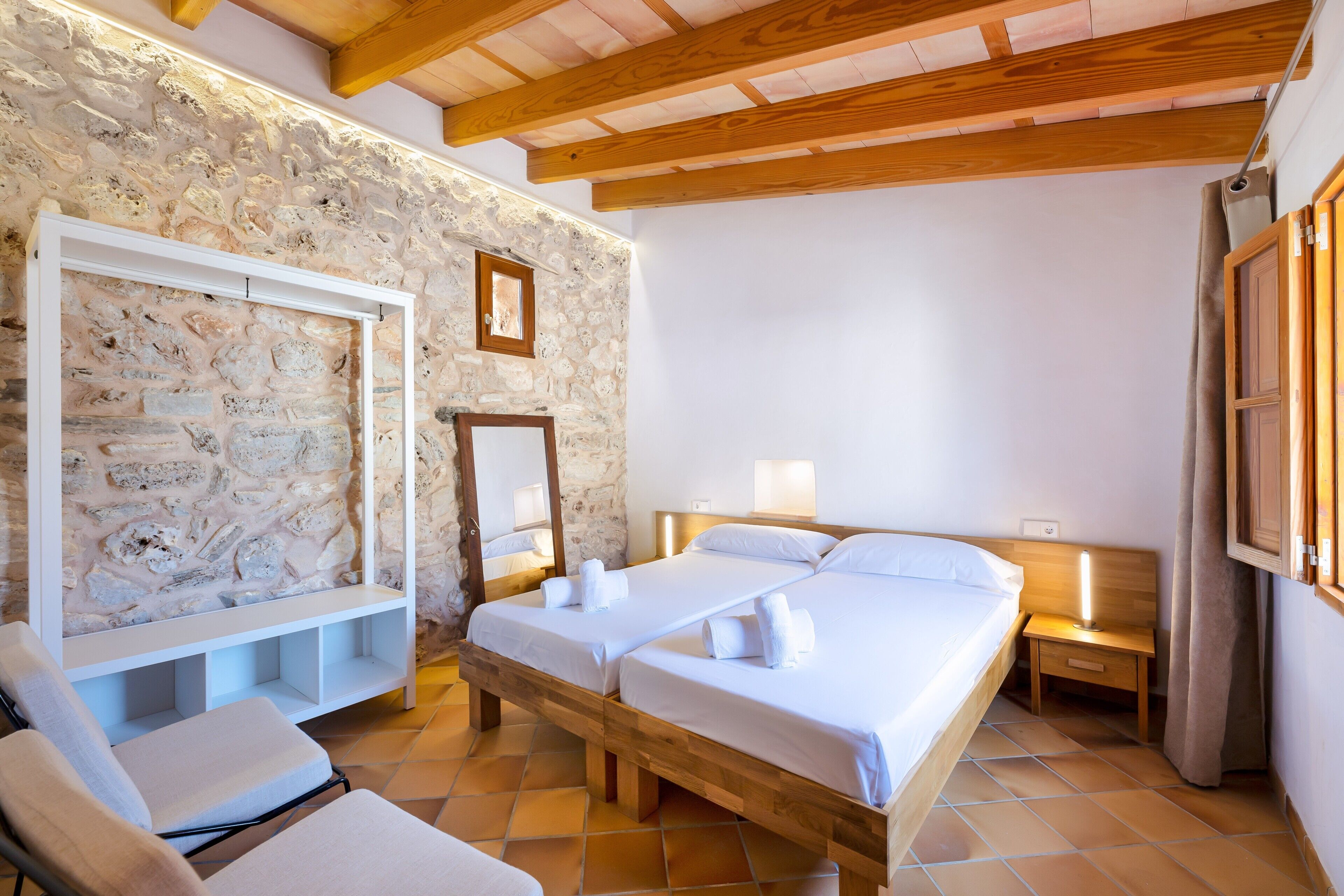 Une chambre avec deux lits, un mur en pierre, des poutres en bois, un banc et une petite table de chevet avec une lampe.