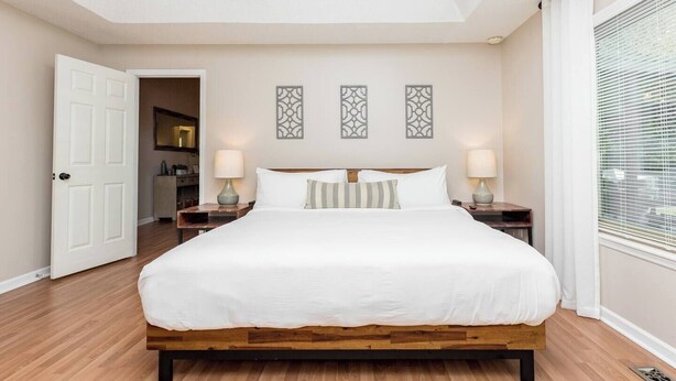 Un dormitorio con una cama grande, dos mesitas de noche, una puerta que da a un baño y una ventana con persianas.
