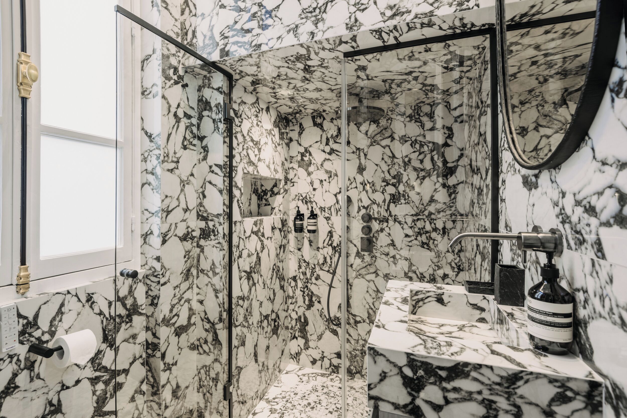 A bathroom with a glass-enclosed shower, marble walls, and a sink with a black soap dispenser.