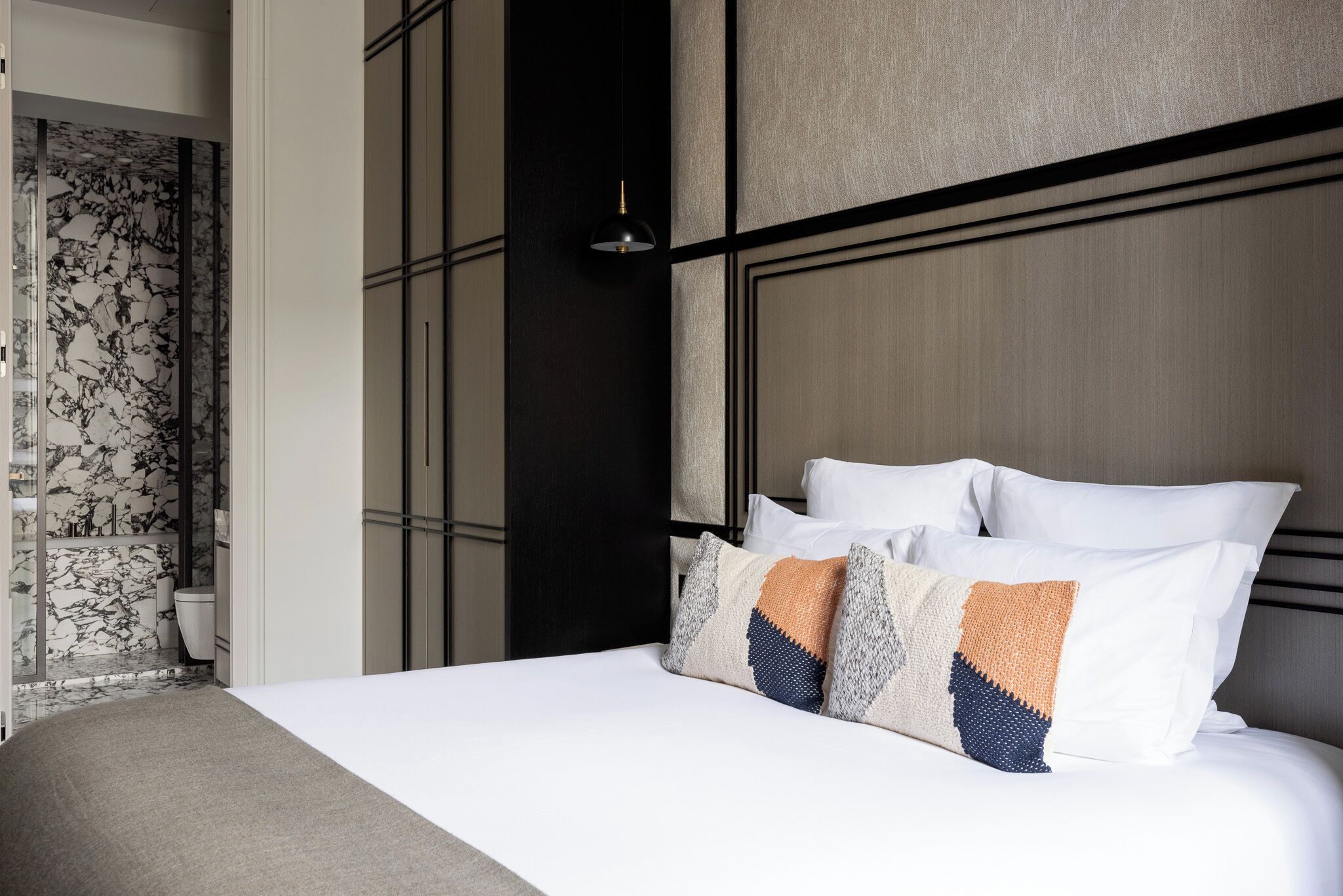A neatly made bed with white and patterned pillows, a dark headboard, and a wall with a geometric design.