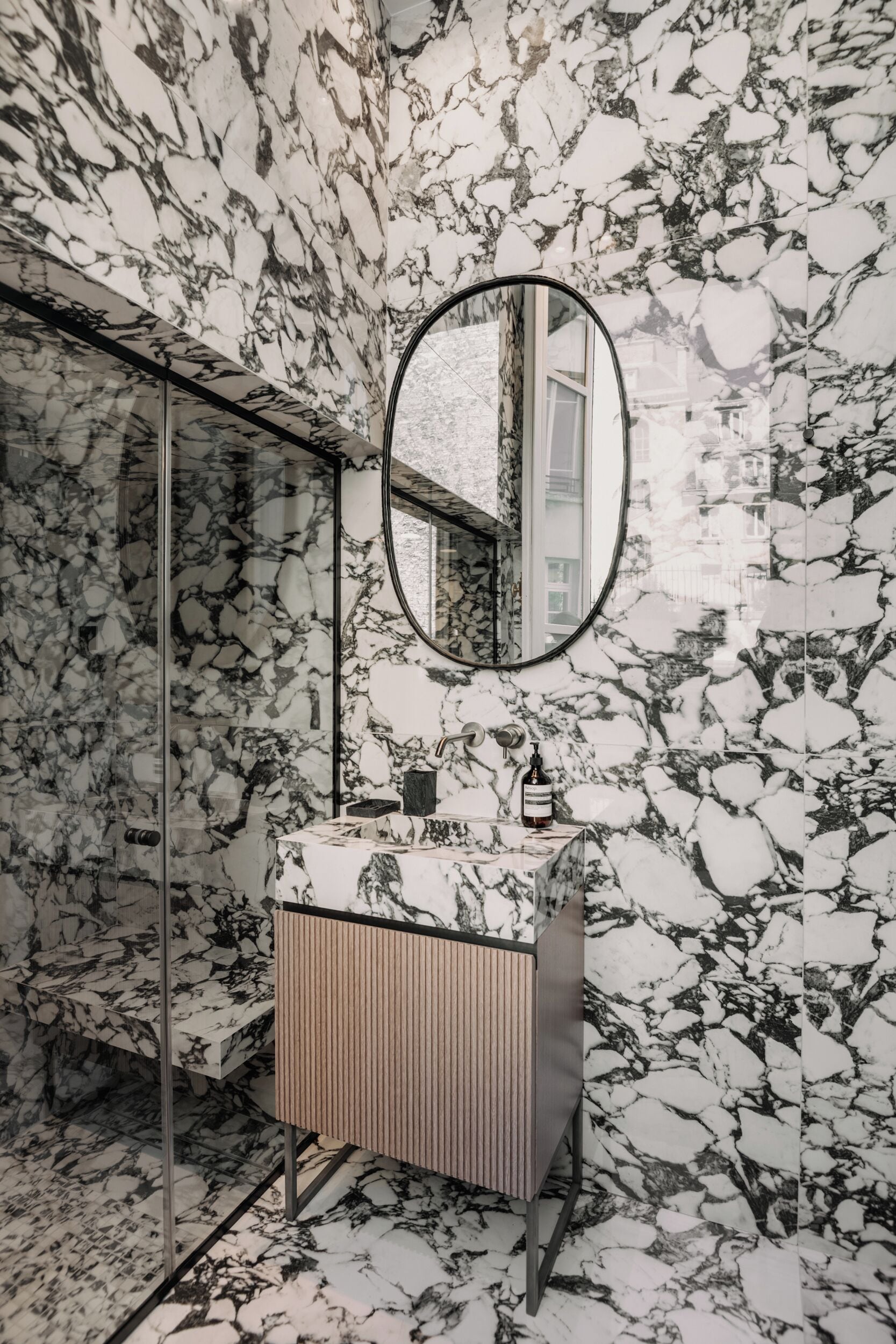 A bathroom with a marble wall, a round mirror, a wooden cabinet, and a glass shower enclosure.