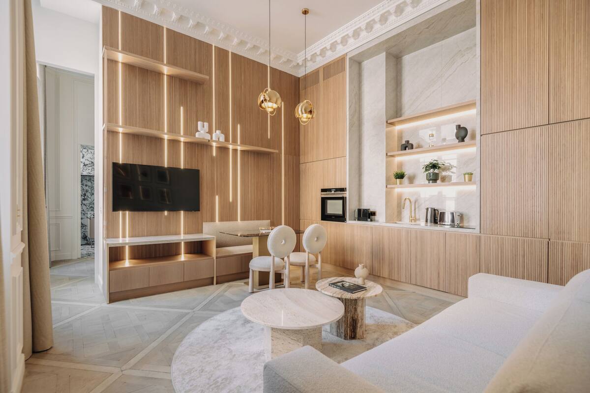 A modern living room with a flat-screen TV, wooden paneling, a sofa, two chairs, a coffee table, and a kitchen area with a sink and countertop.