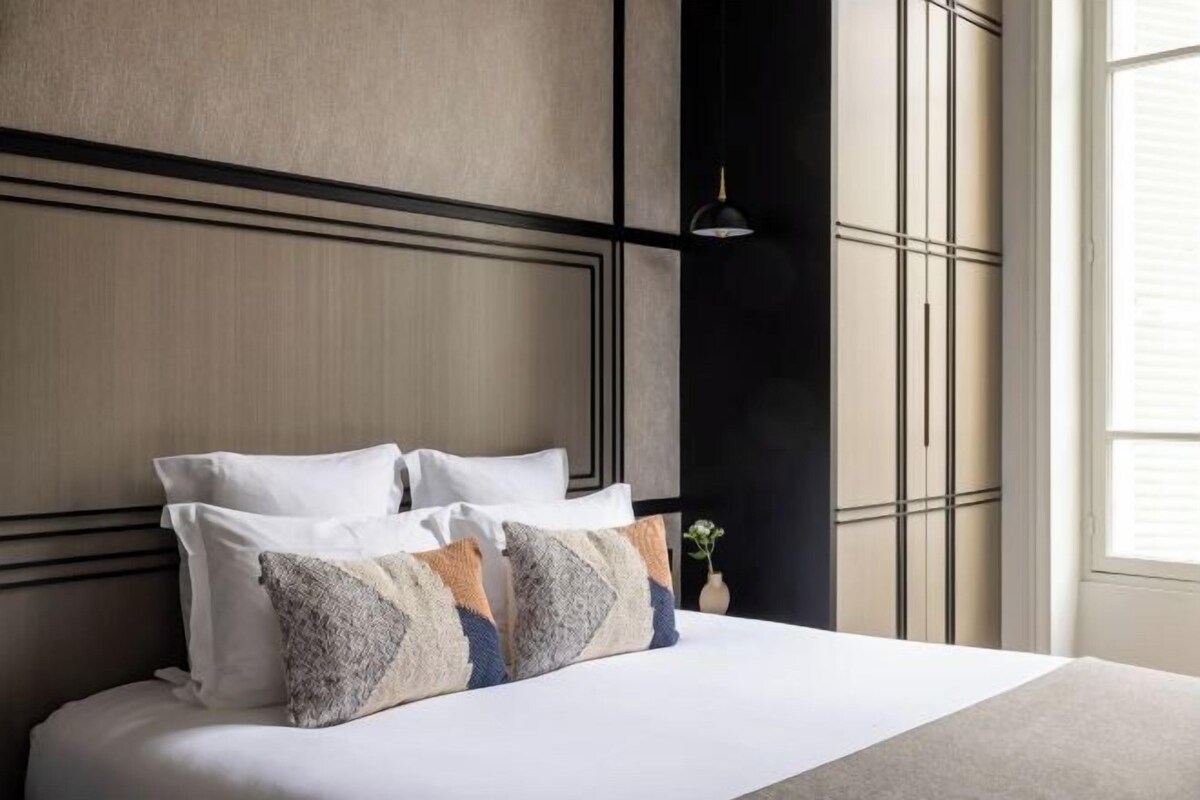 A neatly made bed with white linens and decorative pillows, a modern headboard, and a window with blinds.