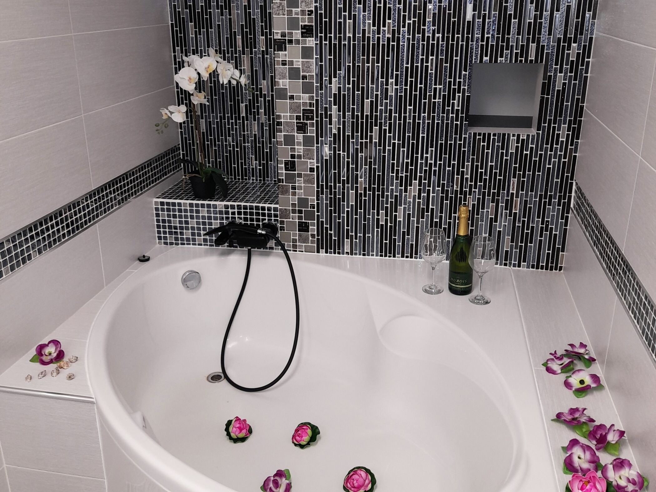 Une salle de bain avec une baignoire blanche, des carreaux de mosaïque noirs et blancs, une plante en pot, une bouteille de champagne et des fleurs décoratives.