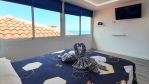 Hotellihuone, jossa on sänky, seinälle asennettu televisio ja suuret ikkunat, joista avautuu näkymä merelle.