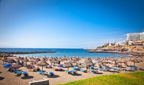 A beach with sun loungers and thatched umbrellas, a rocky shoreline, and a hotel in the background.
