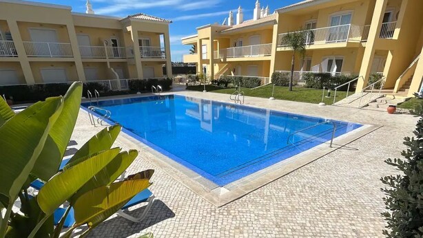 Ein Swimmingpool, umgeben von gelben Gebäuden mit Balkonen, einem strahlend blauen Himmel und einer gefliesten Terrasse.