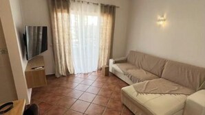 Ein Wohnzimmer mit einem beigen Sofa, einem Wand-Fernseher und einem Fenster mit transparenten Vorhängen.