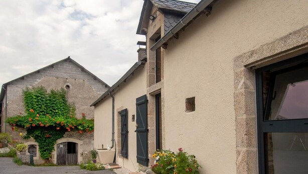 A street with buildings, including a house with a large window and a door, and a stone wall with greenery.
