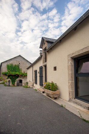 A street with buildings, including a house with a large window and a door, and a stone wall with greenery.